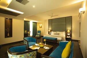 Una habitación de hotel con una cama, dos sillones azules, una mesita de centro con un florero y fruta, y una mesita de noche con una lámpara.