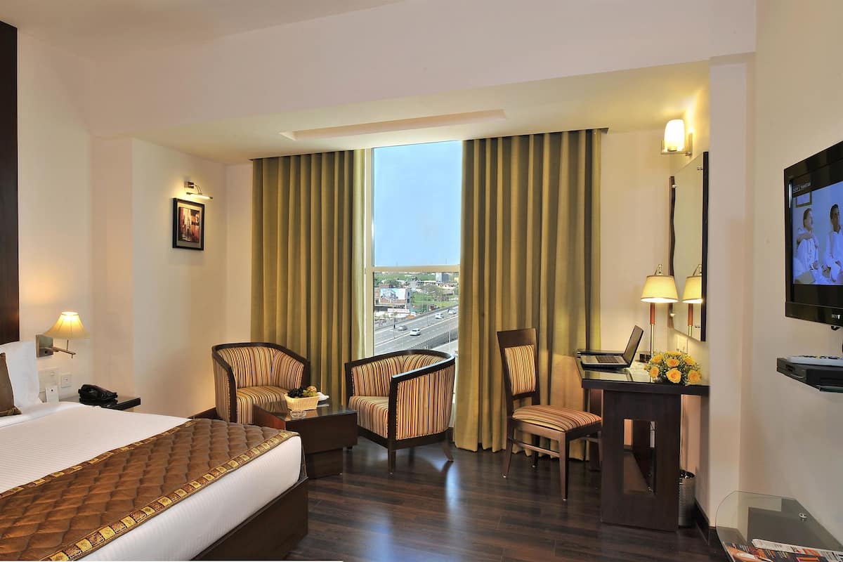A hotel room with a large bed, two armchairs, a desk with a laptop, a TV, and a view of the city through the window.