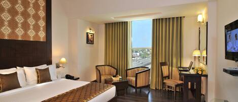 A hotel room with a large bed, two armchairs, a desk with a laptop, a TV, and a view of the city through the window.