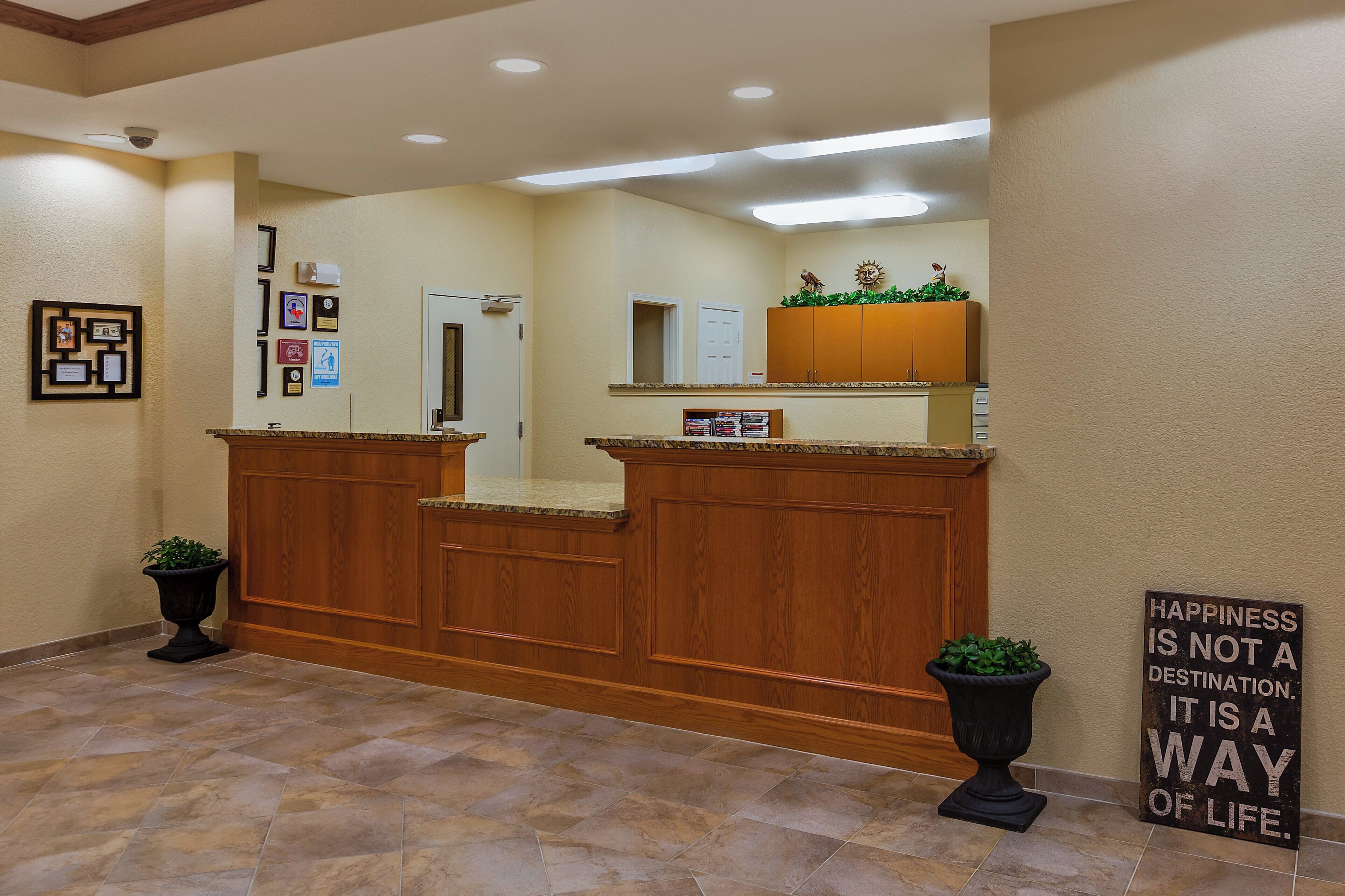 A reception area with a wooden reception desk, a bulletin board with framed photos and notices, a potted plant, and a motivational sign on the wall.
