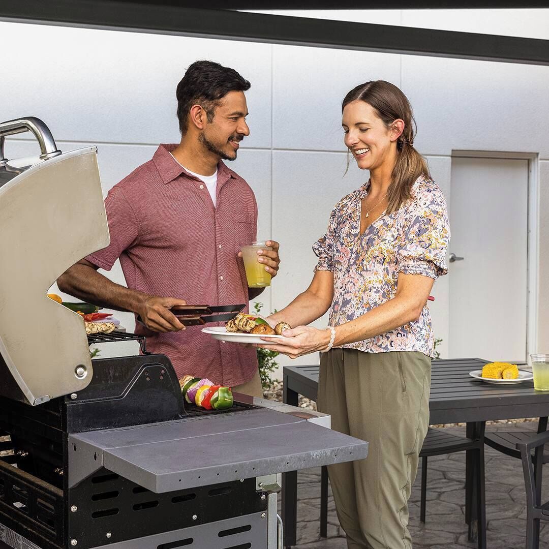 A man grilling food on an outdoor grill with a woman holding a plate of food and a drink.