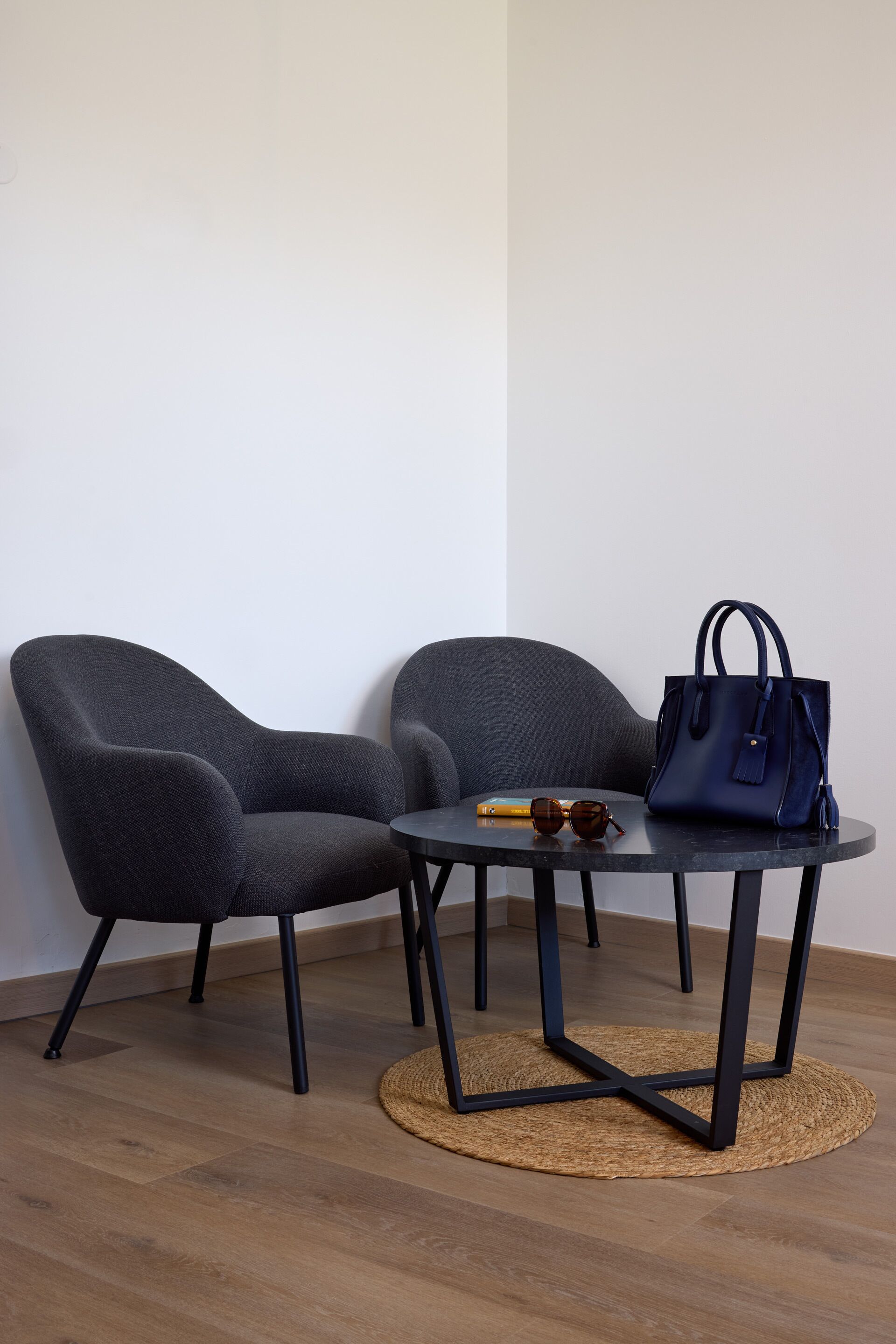 Dos sillones grises, una mesita redonda con un tapete tejido y un bolso azul oscuro.