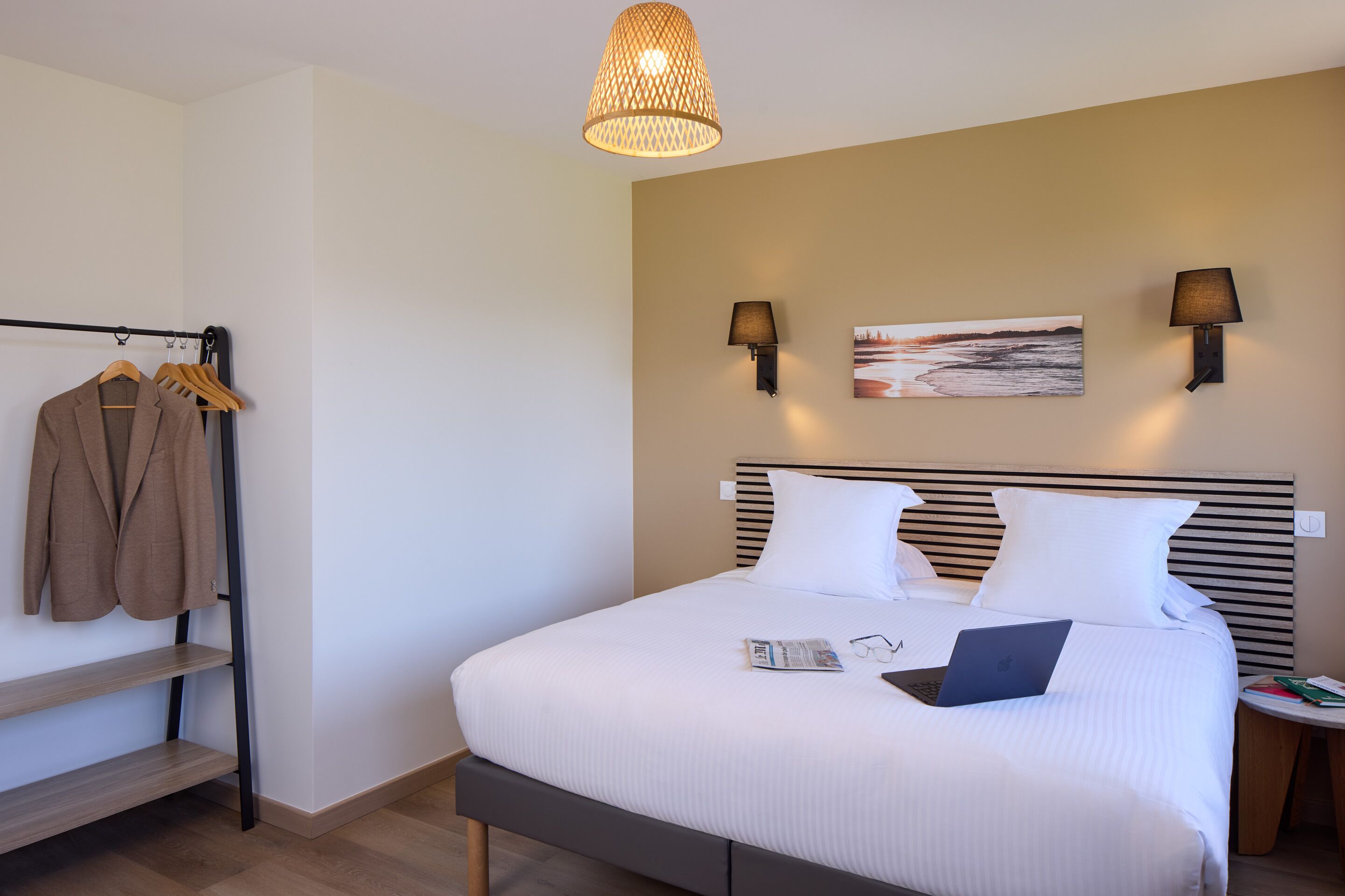 A neatly made bed with white linens, a laptop, and a pair of glasses on it. There are two wall-mounted lamps and a painting on the wall. A wooden shelf with a coat hanger and a pair of shoes is visible.