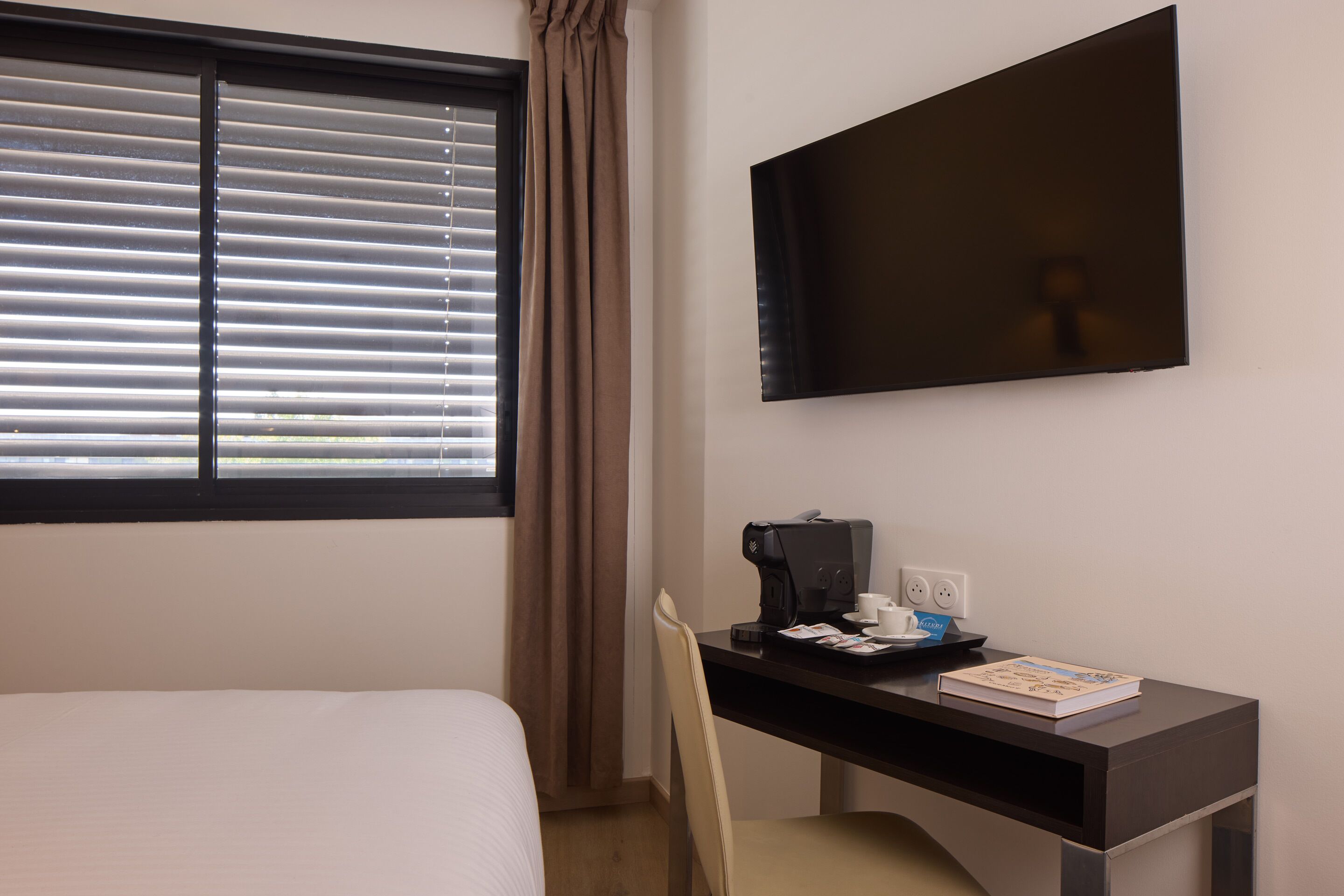 Ein Hotelzimmer mit Bett, Schreibtisch mit Kaffeemaschine, Sessel, Fernseher und Fenster mit Jalousien.