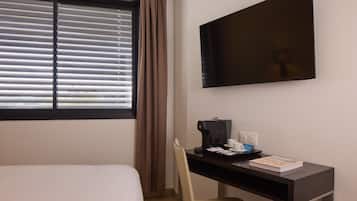 A hotel room with a bed, a desk with a coffee maker, a chair, a TV, and a window with blinds.