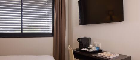 Ein Hotelzimmer mit Bett, Schreibtisch mit Kaffeemaschine, Sessel, Fernseher und Fenster mit Jalousien.