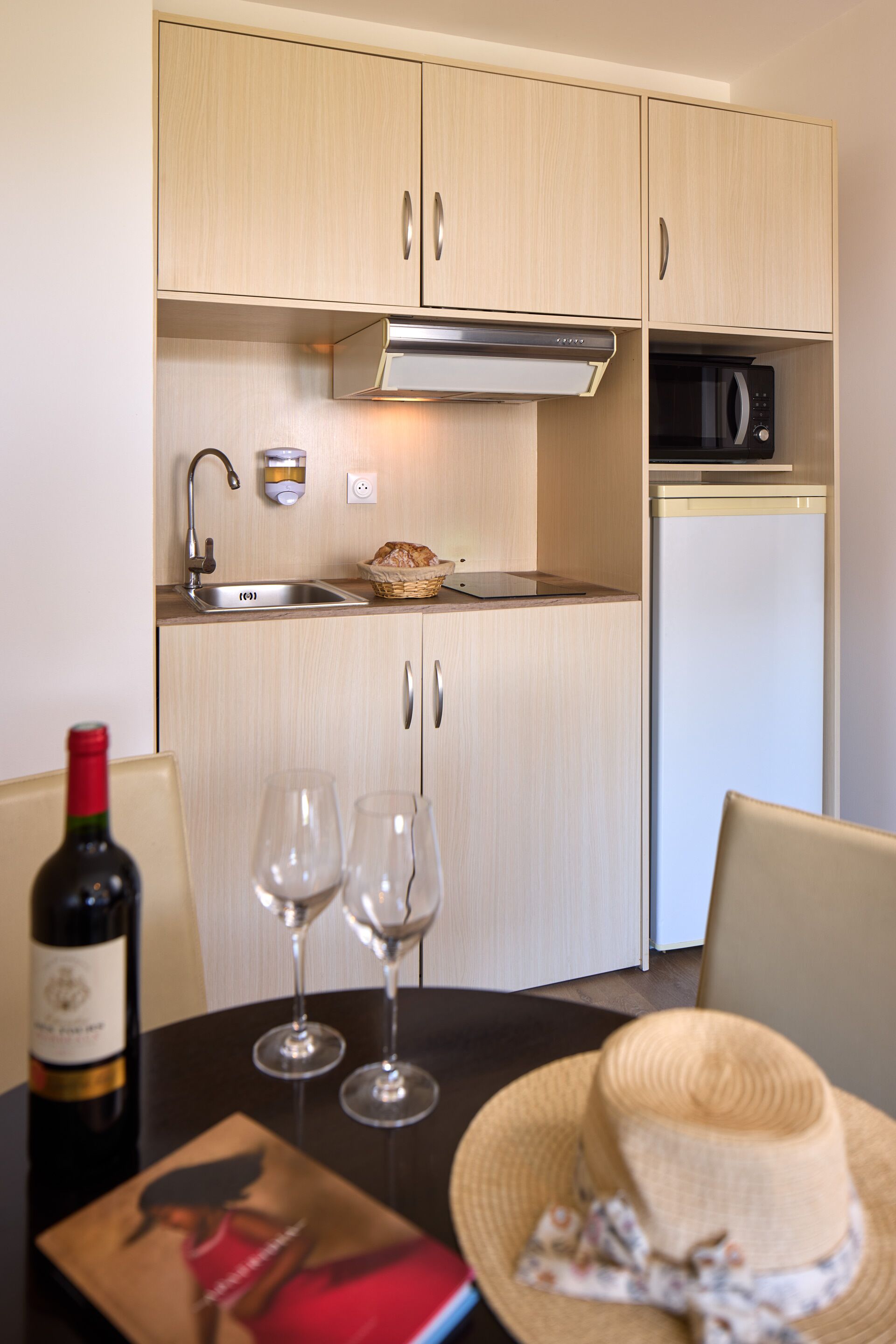 A compact kitchen with wooden cabinets, a microwave, a sink, and a dining table with wine glasses and a bottle.