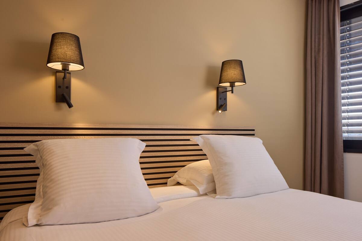 Een hotelkamer met een bed, twee wandlampen en een raam met zonwering.