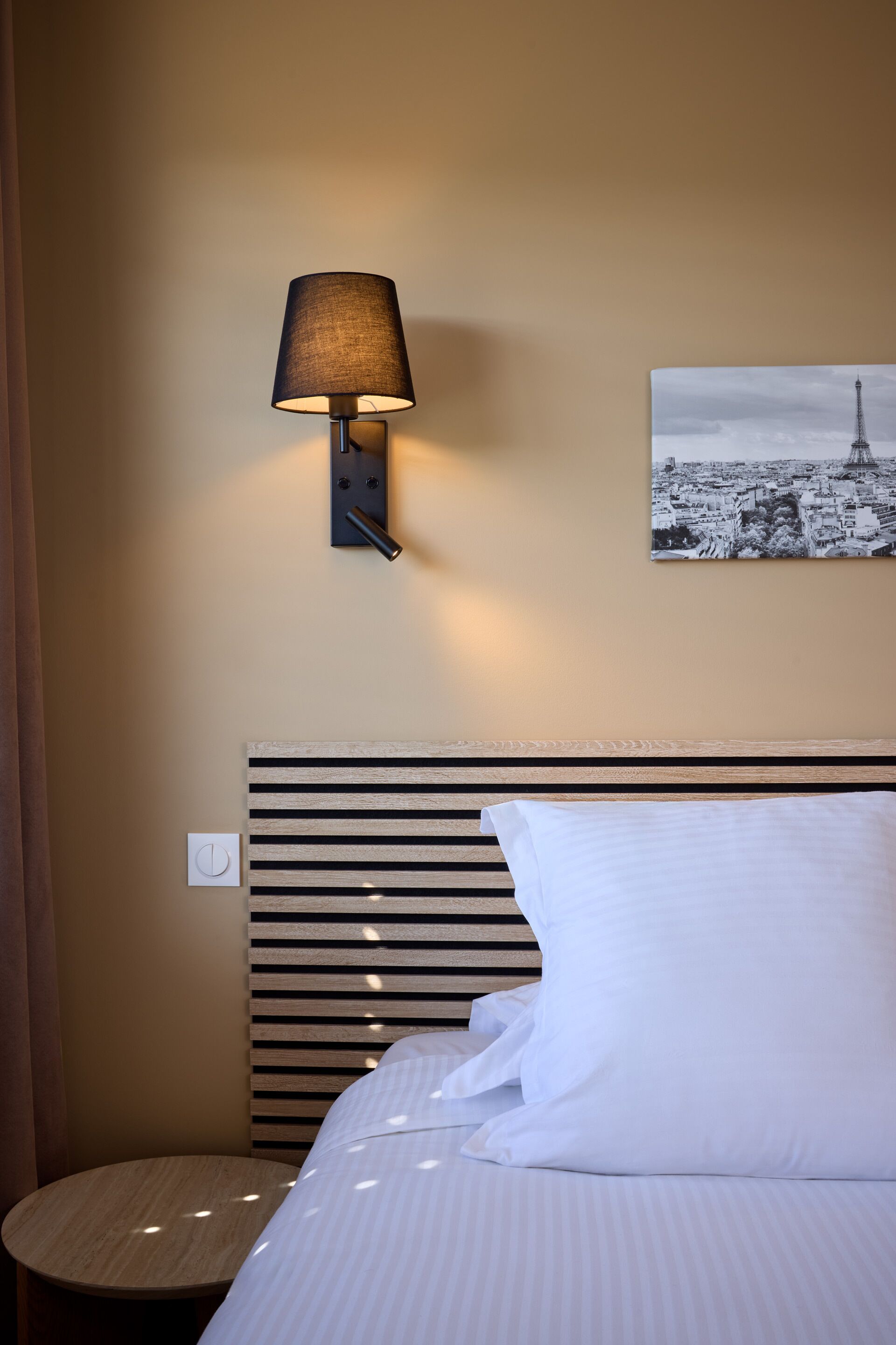 A hotel room with a bed, a wall-mounted lamp, a picture of the Eiffel Tower, and a round wooden table.