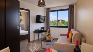 Une chambre d’hôtel avec un lit, un bureau avec un ordinateur, une télévision, une fenêtre avec des rideaux, un canapé avec des coussins et une bouteille de vin sur la table.