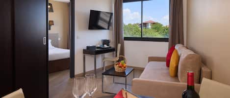 Een hotelkamer met een bed, een bureau met een computer, een televisie, een raam met gordijnen, een bank met kussens, en een fles wijn op de tafel.