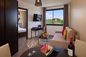 A hotel room with a bed, a desk with a computer, a TV, a window with curtains, a sofa with cushions, and a bottle of wine on the table. - Zenitude Swiss Apparthotels (Ferney-Voltaire)