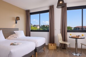 A hotel room with two beds, a dining table with chess, a chair, and a view of buildings. - Zenitude Swiss Apparthotels (Ferney-Voltaire)