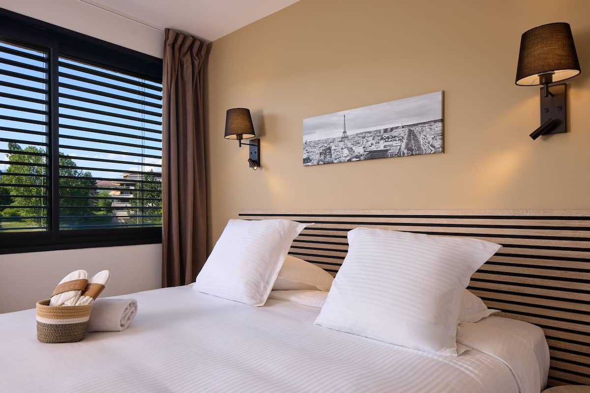 Hotelkamer met een bed, twee kussens, een nachtkastje met een lamp, een schilderij aan de muur en een raam met jaloezieën.