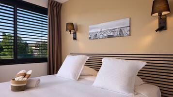 Habitación de hotel con una cama, dos almohadas, una mesita de noche con una lámpara, un cuadro en la pared y una ventana con persianas.