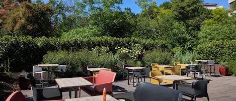 An outdoor patio with tables and chairs, surrounded by greenery and trees.