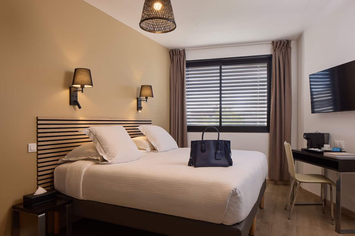 Een hotelkamer met een bed, nachtlampjes, een bureau met stoel, een televisie en een raam met zonwering.