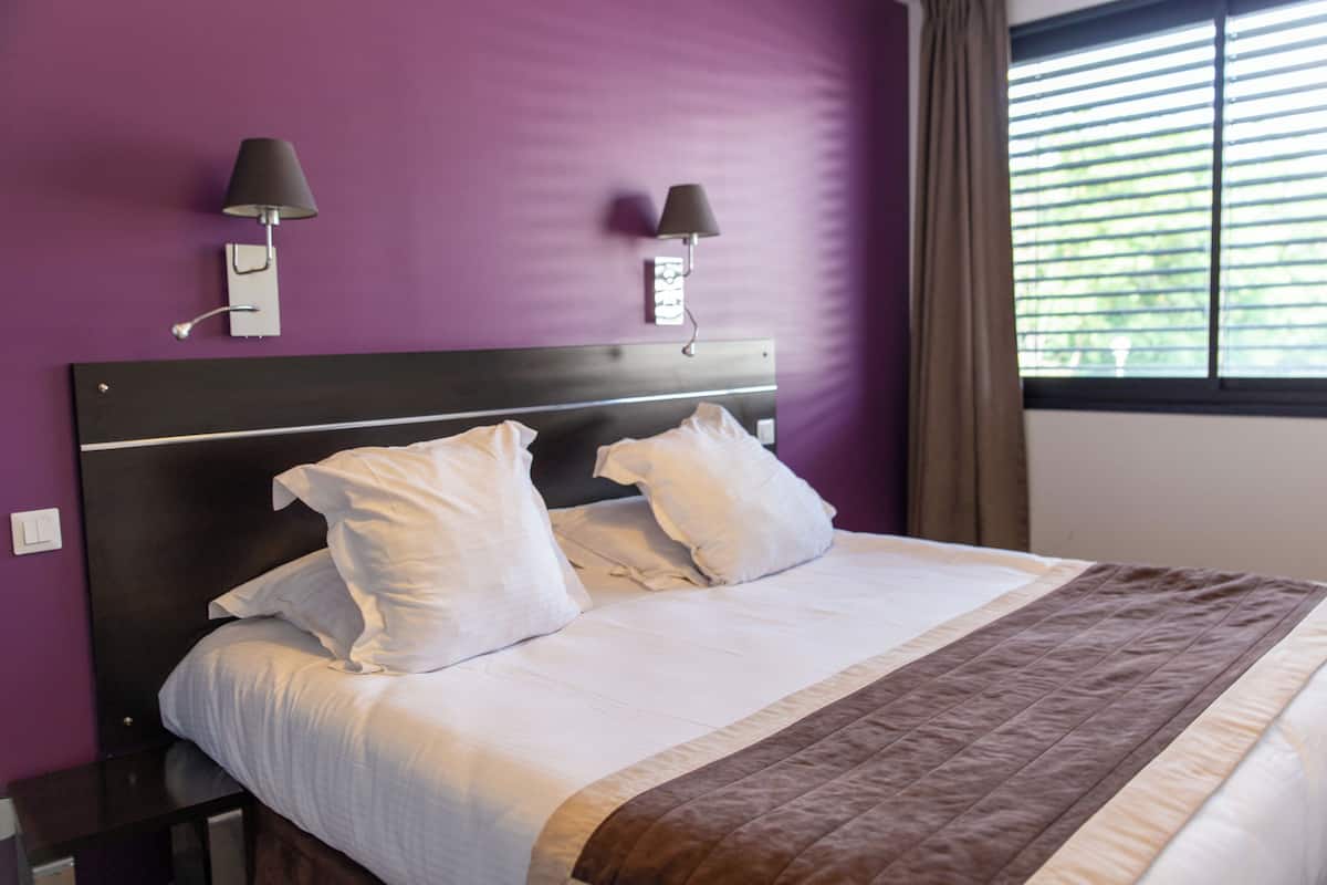 Een net opgemaakt bed met witte lakens en een bruine plaid, een donker hoofdbord en twee wandlampen.