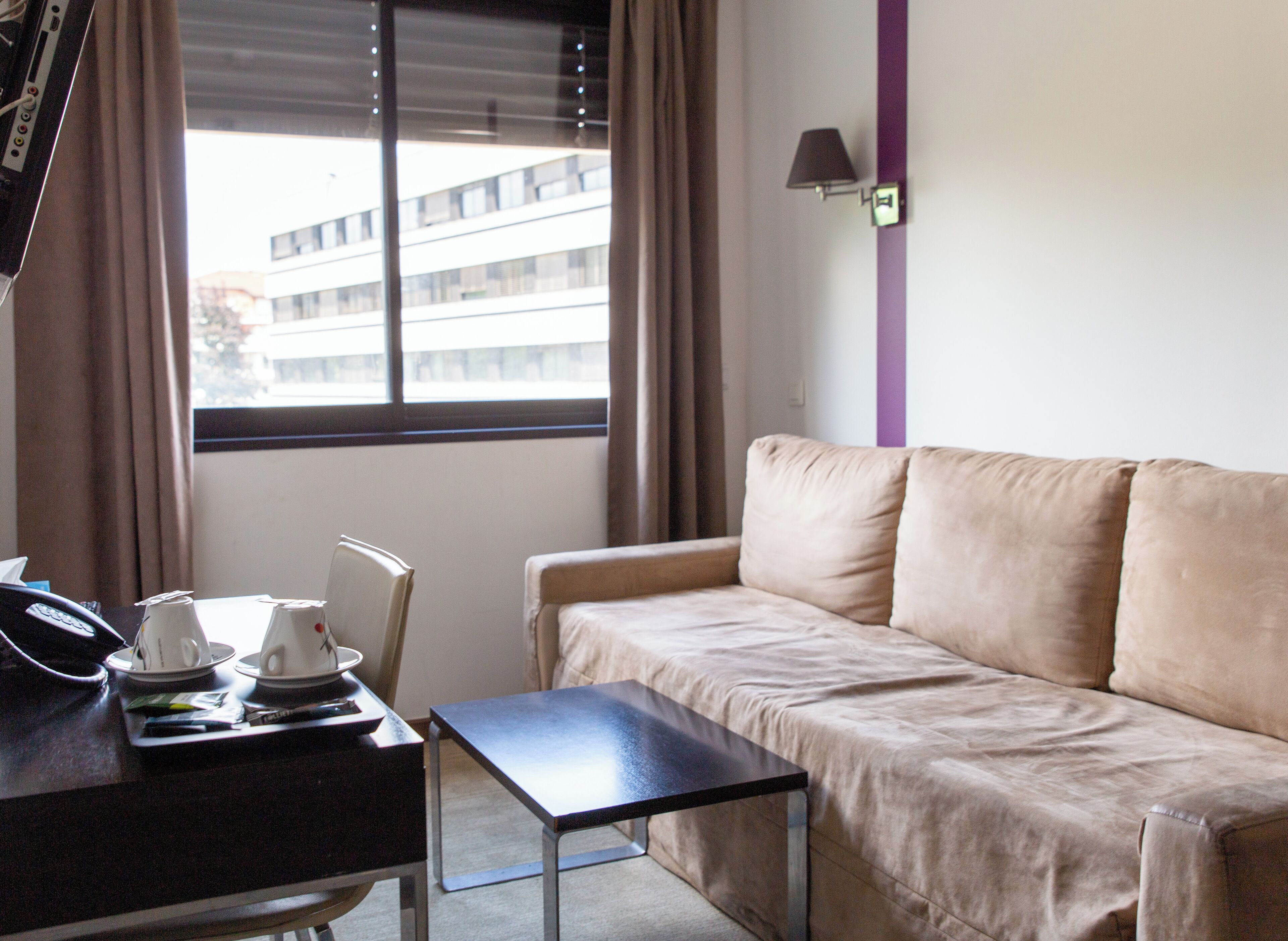 Una habitación de hotel con sofá, una mesita con teléfono y tazas, y una ventana con persianas.