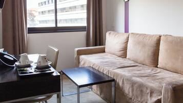 Een hotelkamer met een bank, een kleine tafel met een telefoon en bekers, en een raam met jaloezieën.