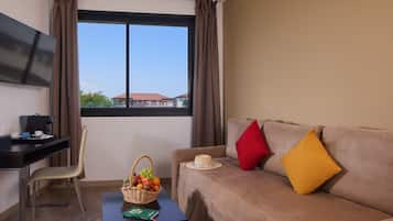 Una habitación con sofá, escritorio, televisor, ventana con cortinas y una cesta de fruta sobre una mesita.