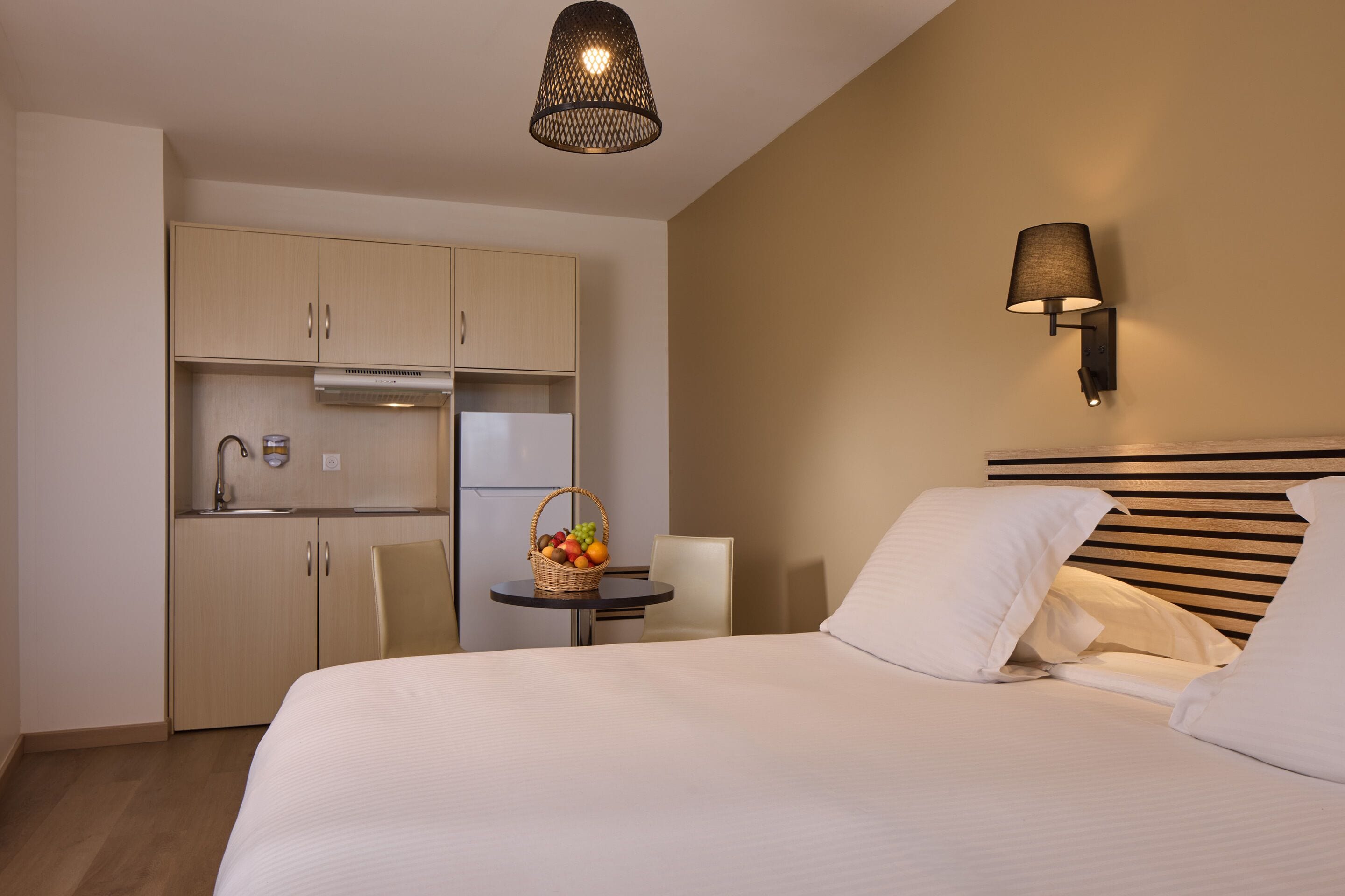 Une chambre d’hôtel avec un lit, une petite table sur laquelle se trouve un panier de fruits, un coin cuisine et une lampe fixée au mur.