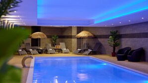 Une piscine entourée de murs, agrémentée de chaises longues et de parasols, et dont le plafond est illuminé par des luminaires.