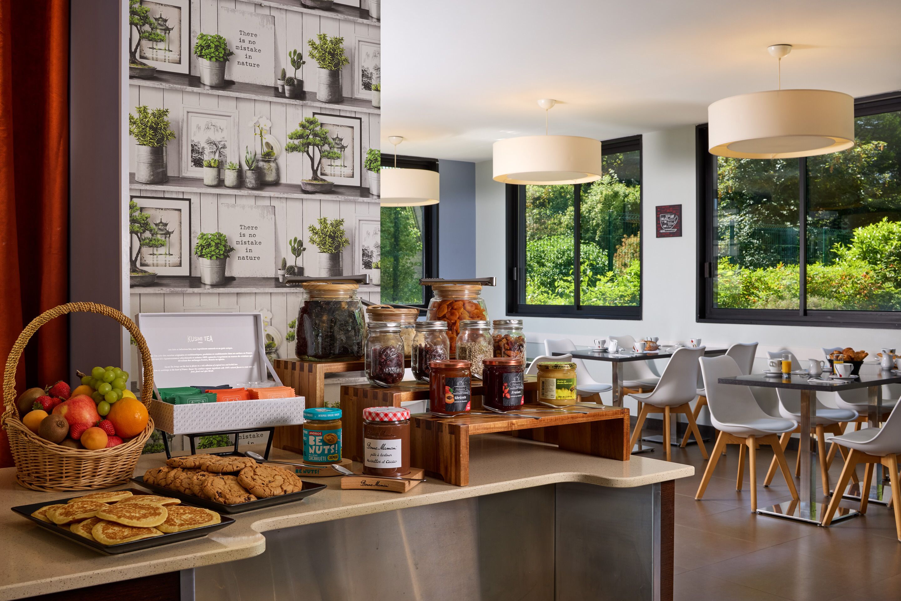 A breakfast buffet with various food items, a wall with framed pictures, and a view of greenery through large windows.