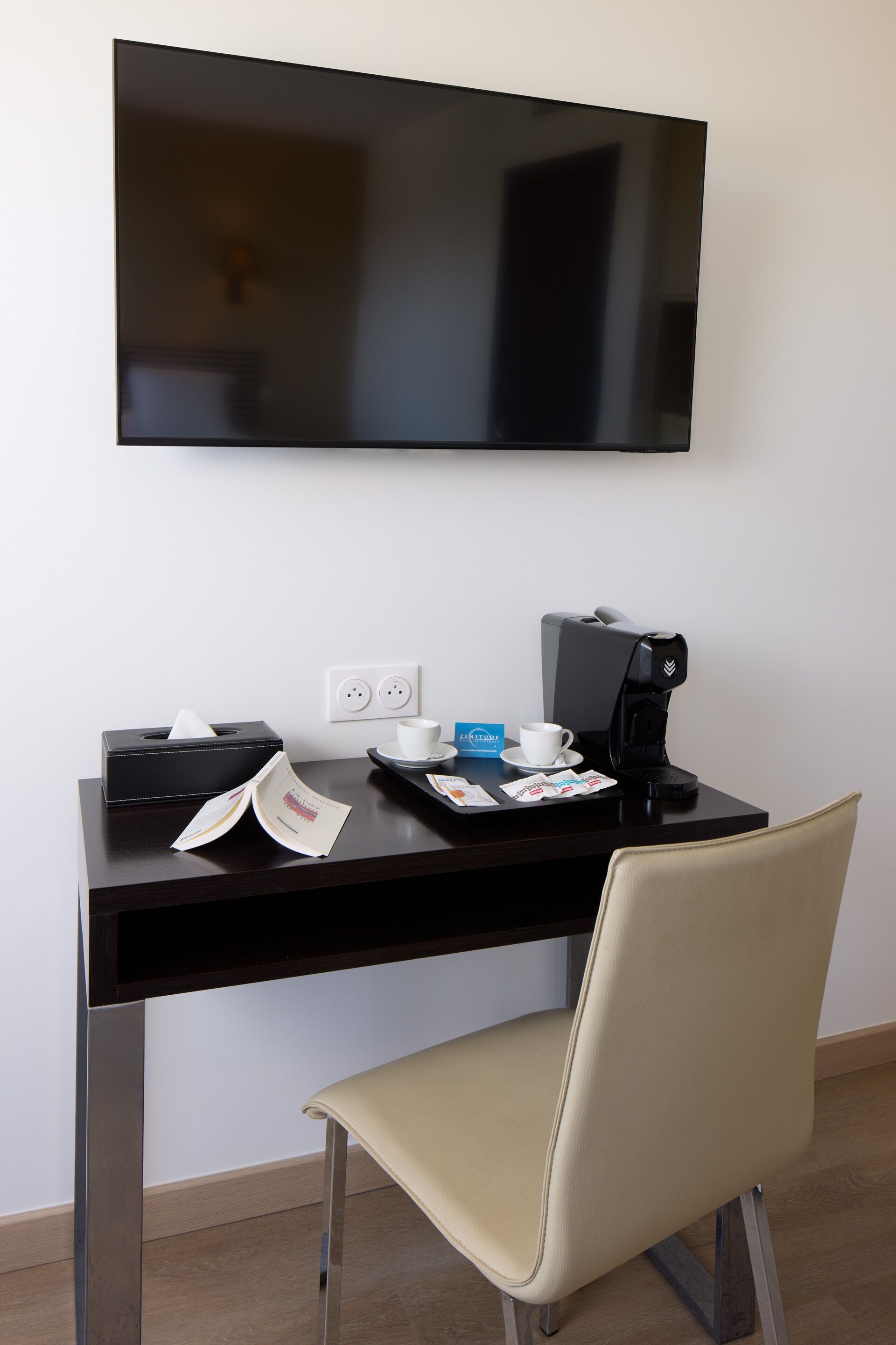 Ein Hotelzimmer mit Schreibtisch, Stuhl, Kaffeemaschine, Taschentuchbox, Tasse und einem an der Wand befestigten Flachbildfernseher.
