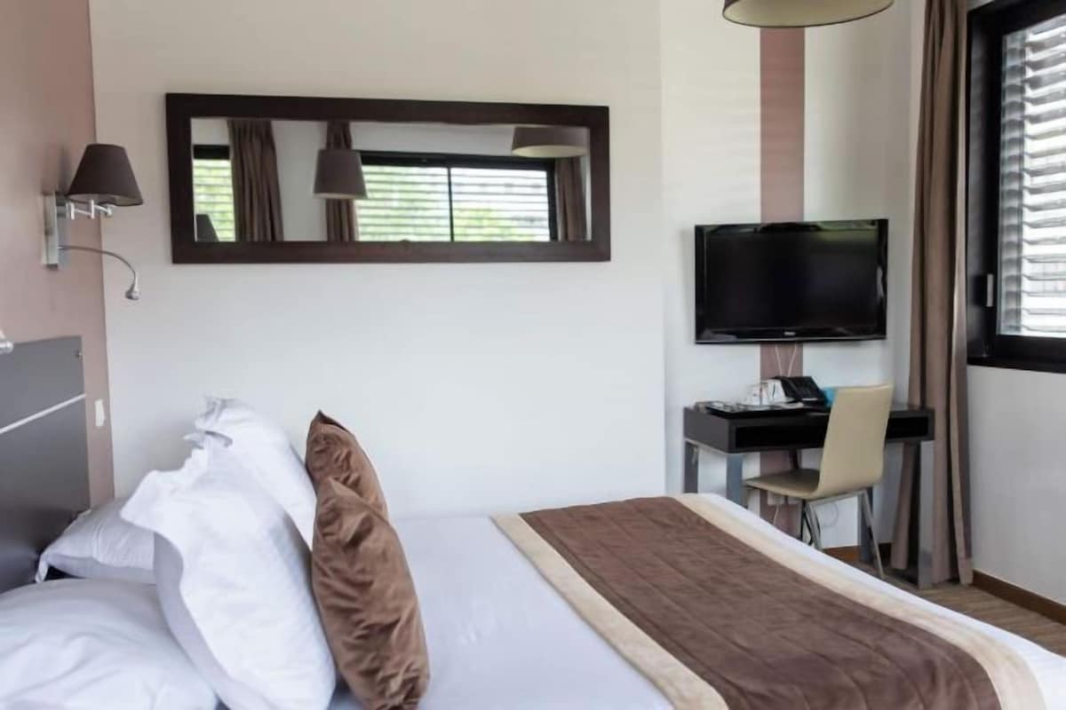 A hotel room with a bed, a desk with a chair, a television, and a window with curtains.