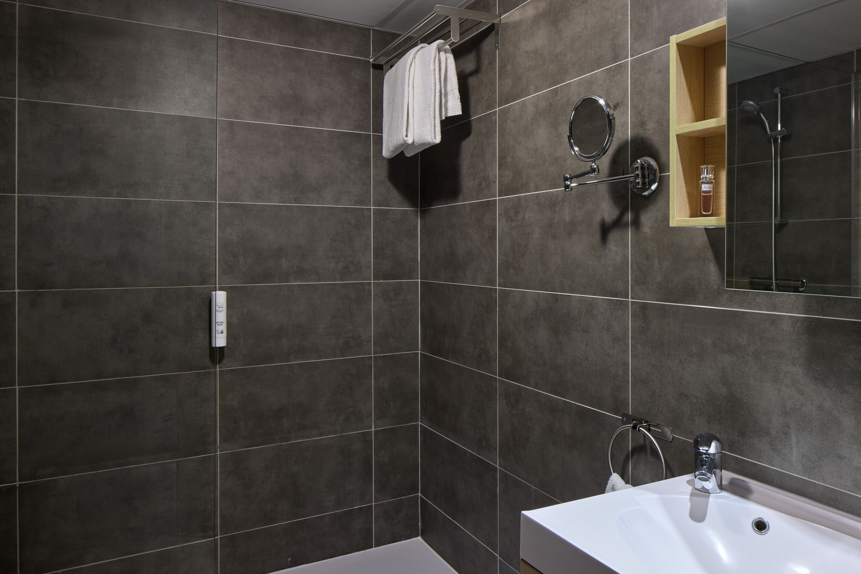 Un baño con baldosas oscuras, un lavabo blanco, un espejo, un perchero para toallas y una ducha.