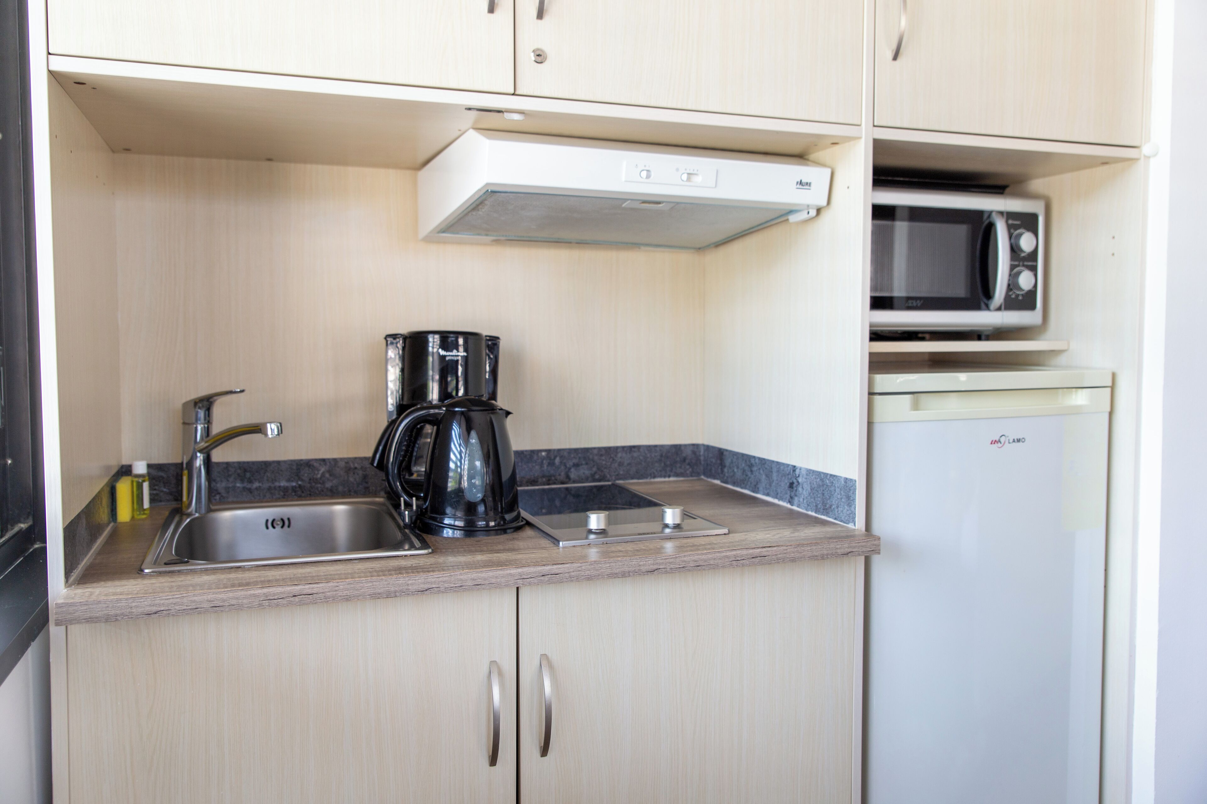Una kitchenette compacta con lavamanos, cafetera, microondas y un refrigerador pequeño.