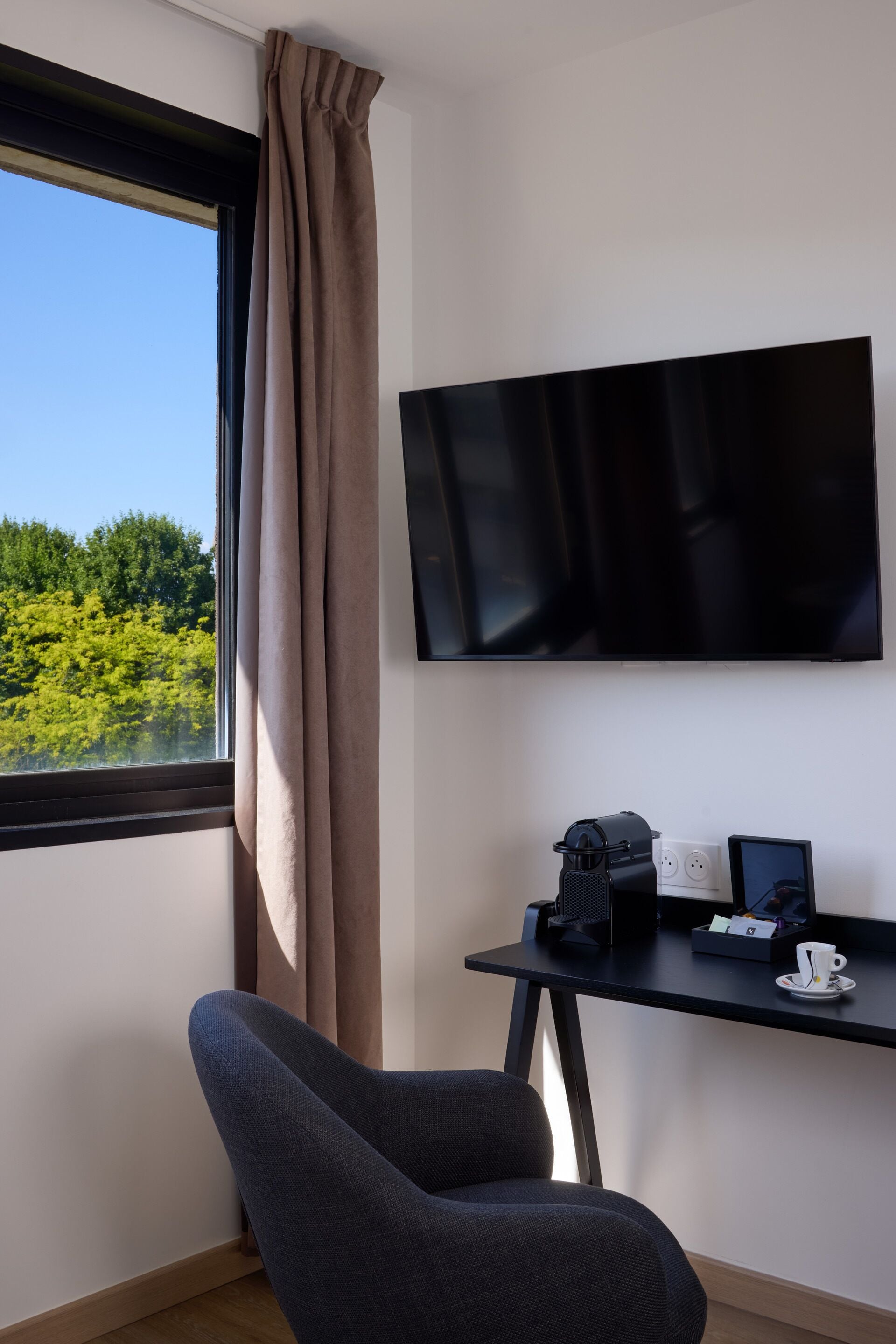 A modern room with a large window, a flat-screen TV, a desk with a coffee maker, and a chair.