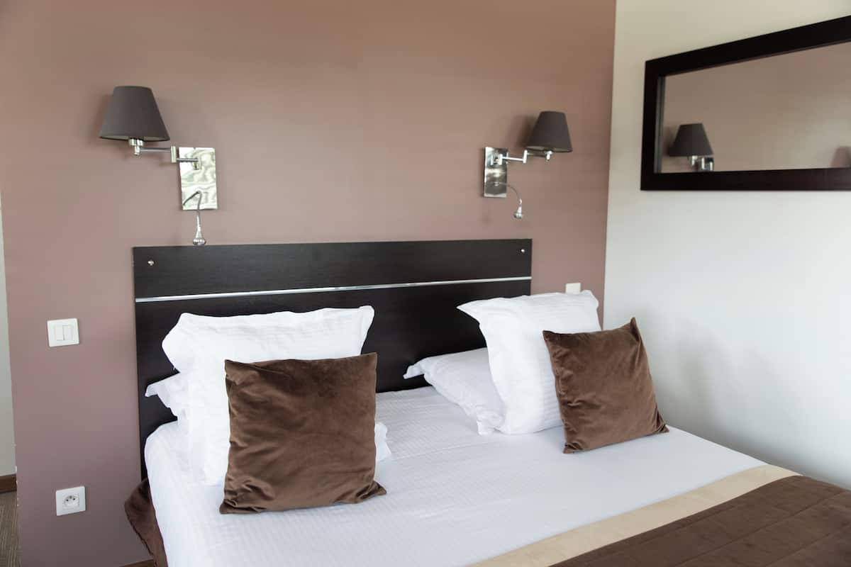 Een keurig opgemaakt bed met witte en bruine kussens, twee wandlampen en een spiegel aan de muur.