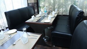 A dining area with a long table set for four, black leather seating, and a view of greenery through sheer curtains. - Hotel Hari Heritage (Haridwar)