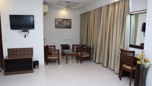 A living room with a flat-screen TV, a sofa, a dining table with chairs, and a large window with curtains. - Hotel Hari Heritage (Haridwar)