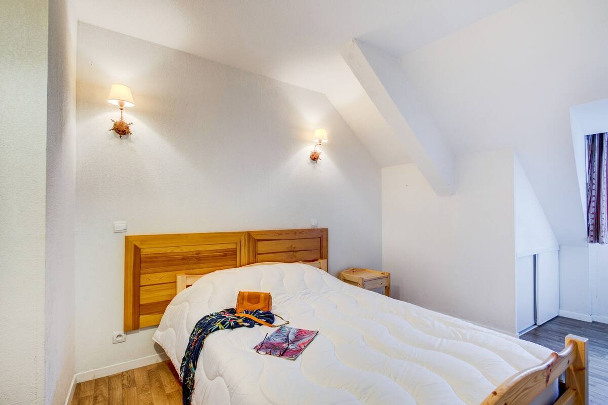 Une chambre à coucher avec une tête de lit en bois, un lit recouvert d’une couette blanche, deux lampes fixées au mur et une petite table de chevet.