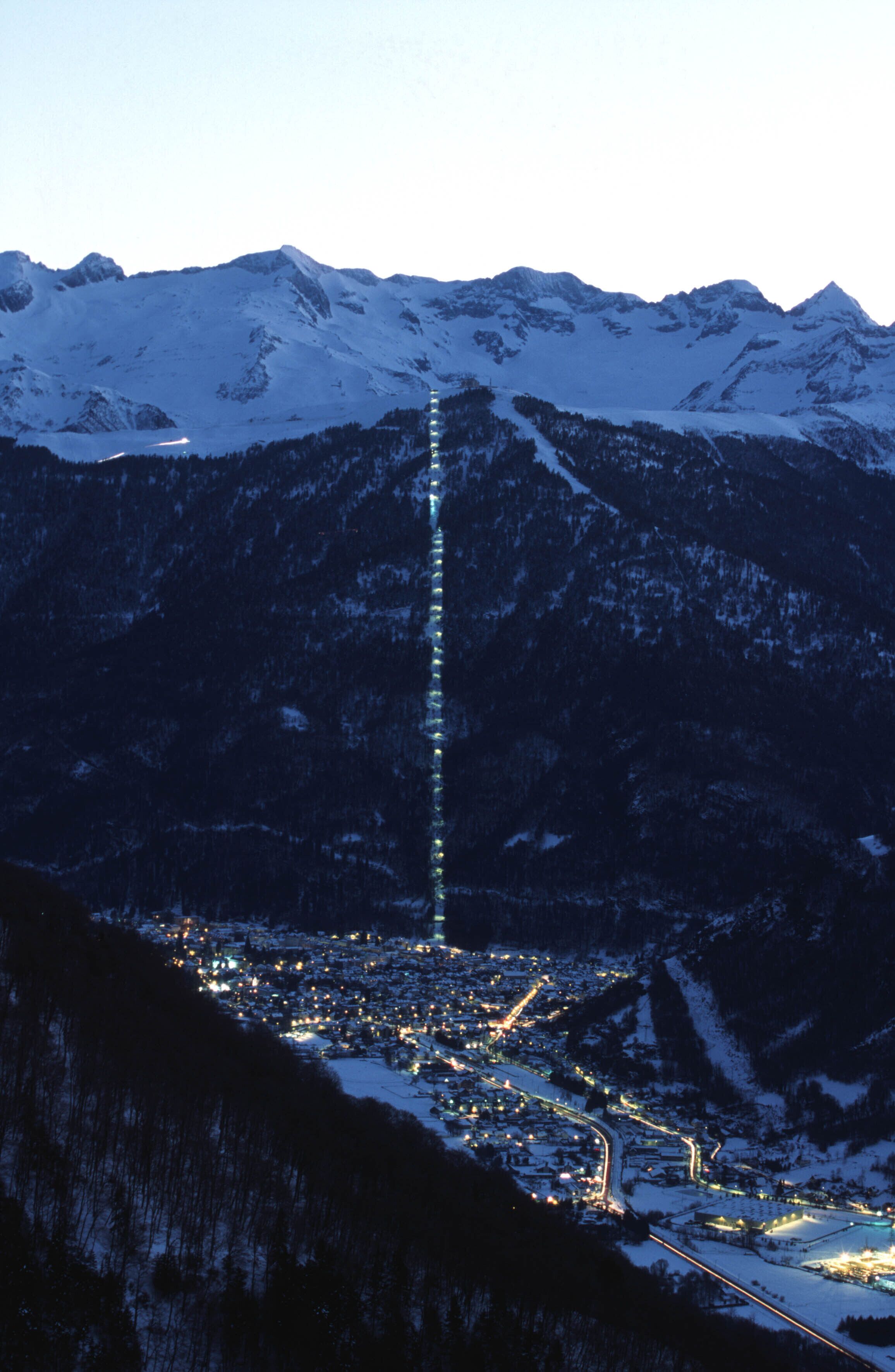 Une chaîne de montagnes enneigées, avec une ville située à son pied, et une structure lumineuse verticale particulièrement visible.