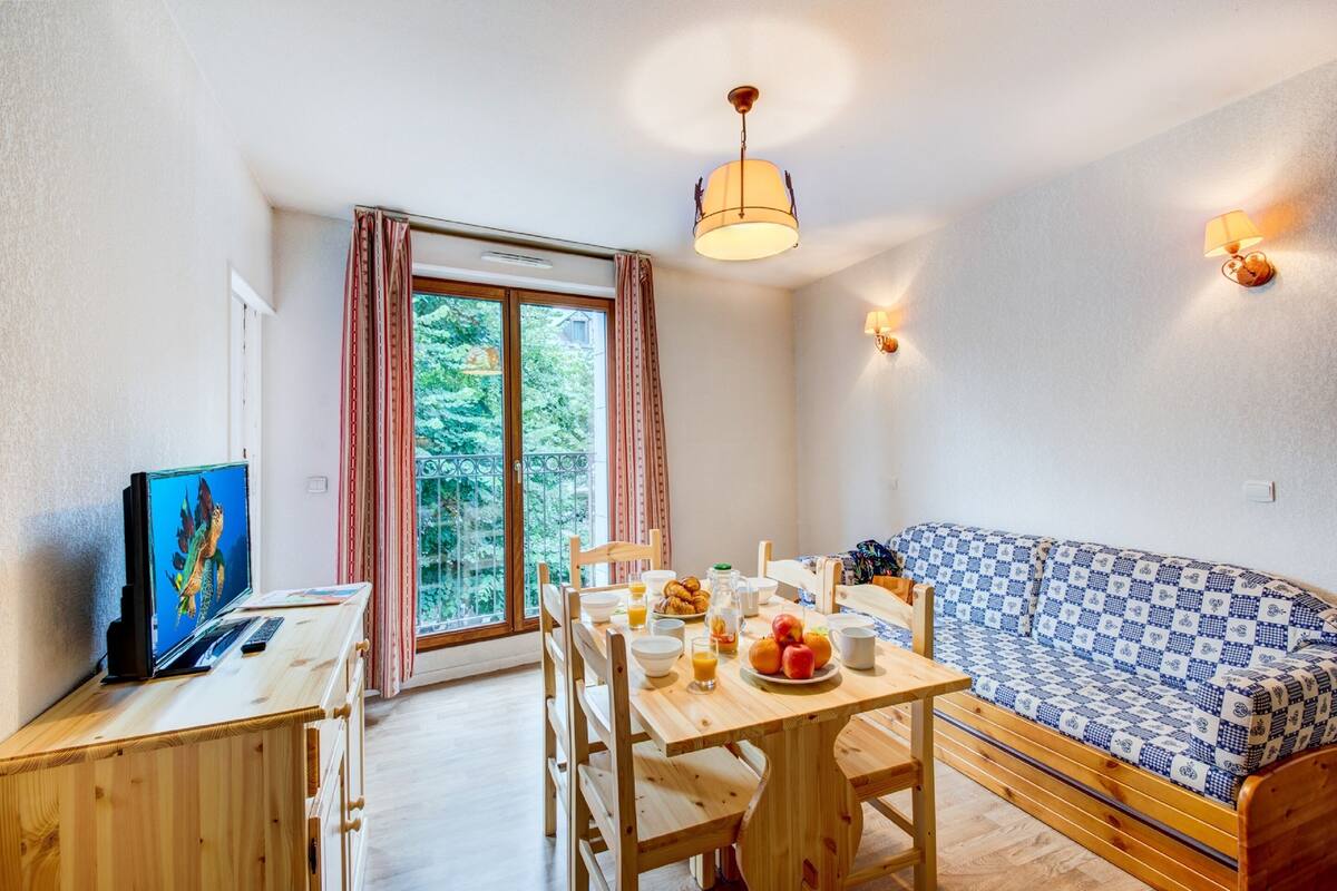Une pièce chaleureuse avec une table à manger en bois, des chaises, un canapé, un téléviseur et une grande fenêtre avec des rideaux.
