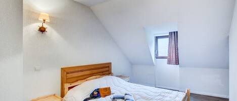 Un dormitorio con techo inclinado, una cama de madera, una mesita de noche, una lámpara de pared y una ventana con cortinas.