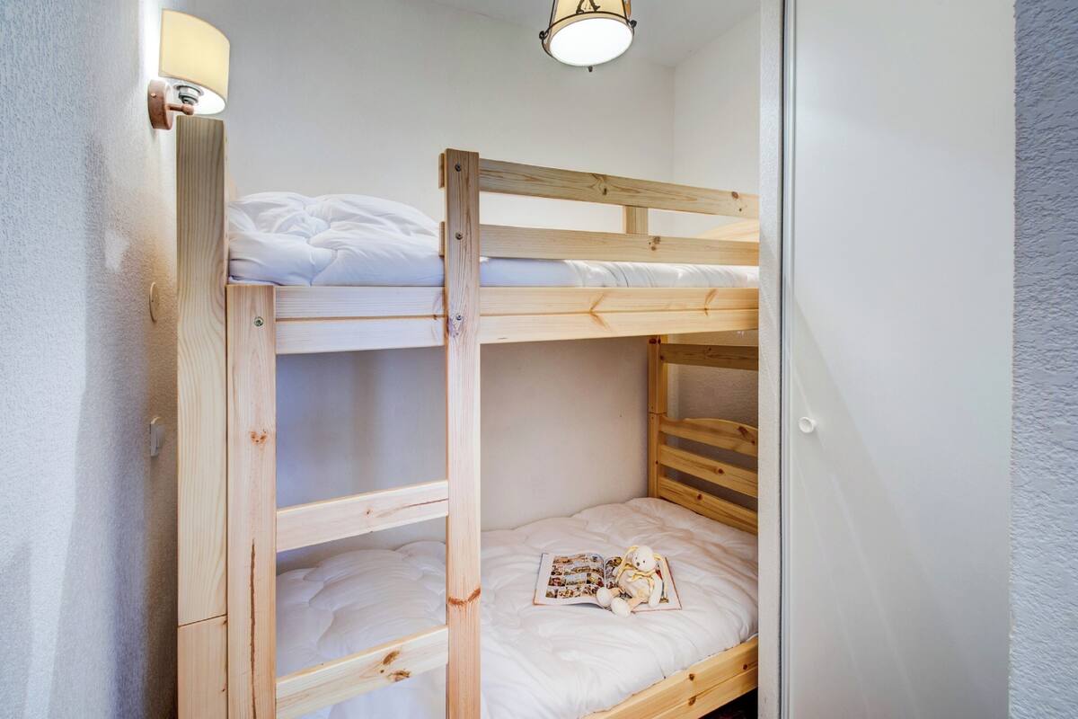 Un lit superposé avec une structure en bois, une literie blanche et une petite table sur laquelle se trouvent un livre et un ours en peluche.