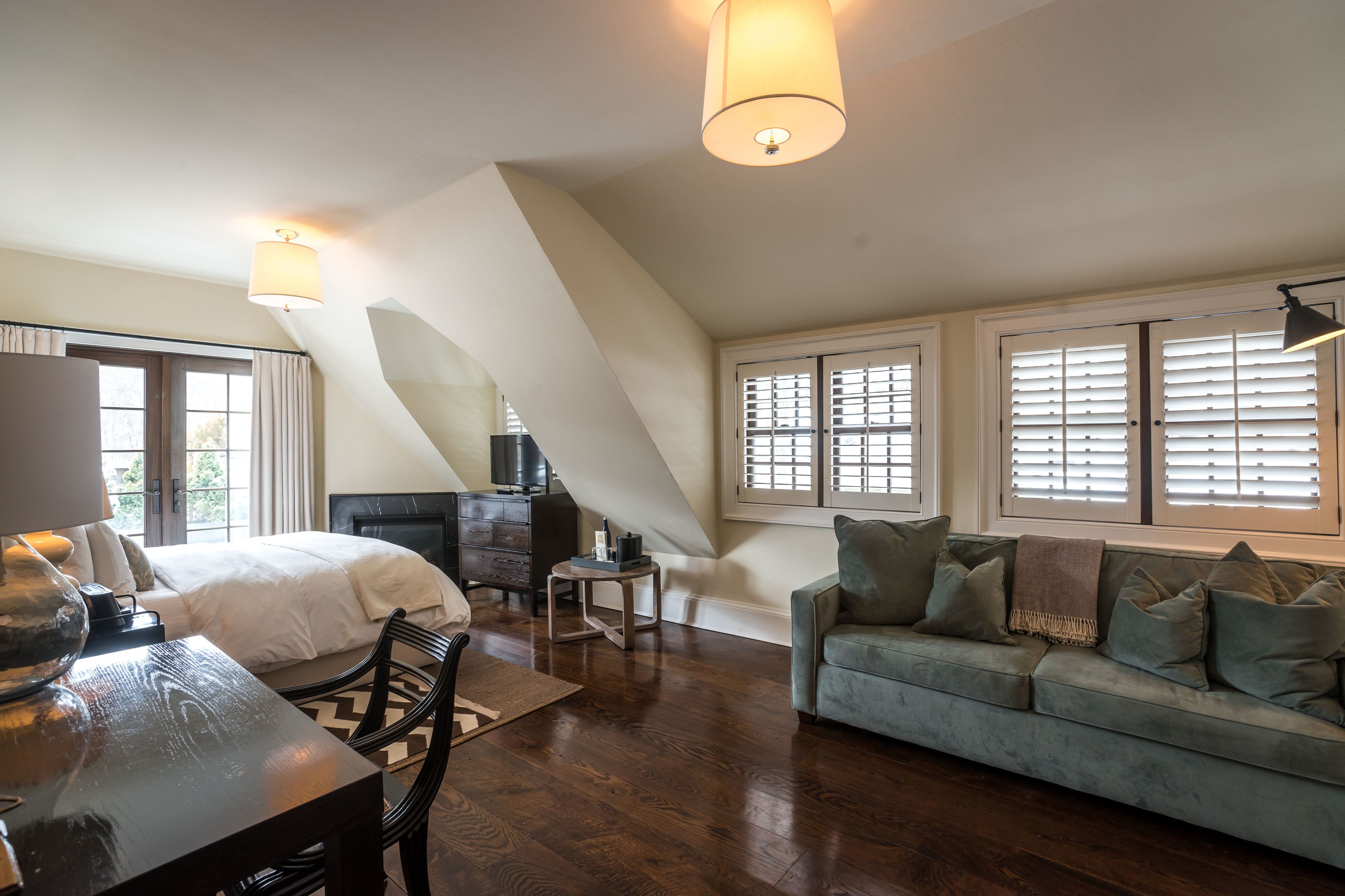Ein geräumiges Schlafzimmer im Loft-Stil mit einem großen Bett, einem Schreibtisch und einer Couch. Es gibt Fenster mit Jalousien, einen Kamin und eine Deckenleuchte.