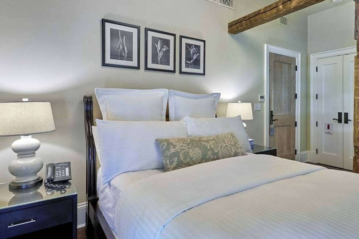 A neatly made bed with white linens and decorative pillows, a bedside table with a lamp and phone, and framed botanical prints on the wall.