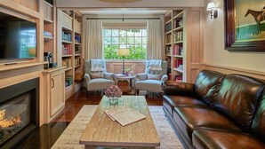 A cozy living room with a fireplace, leather sofa, and a coffee table with a book. There are two armchairs and a bookshelf filled with books. - Bedford Post Inn (Bedford)