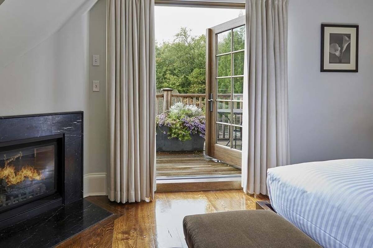 A bedroom with a fireplace, a bed, a balcony with plants, and a view of the outdoors.
