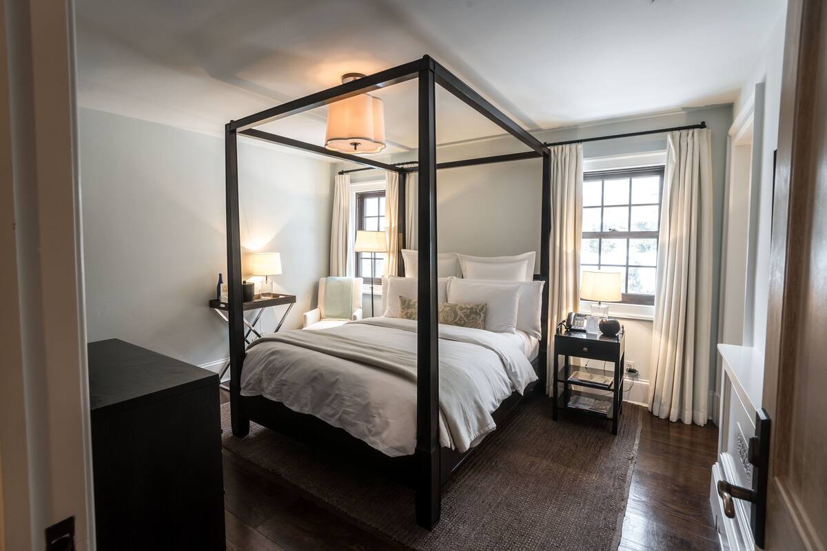 A four-poster bed with a dark frame, a bedside table with a lamp, and a view of the outdoors through a window with curtains.