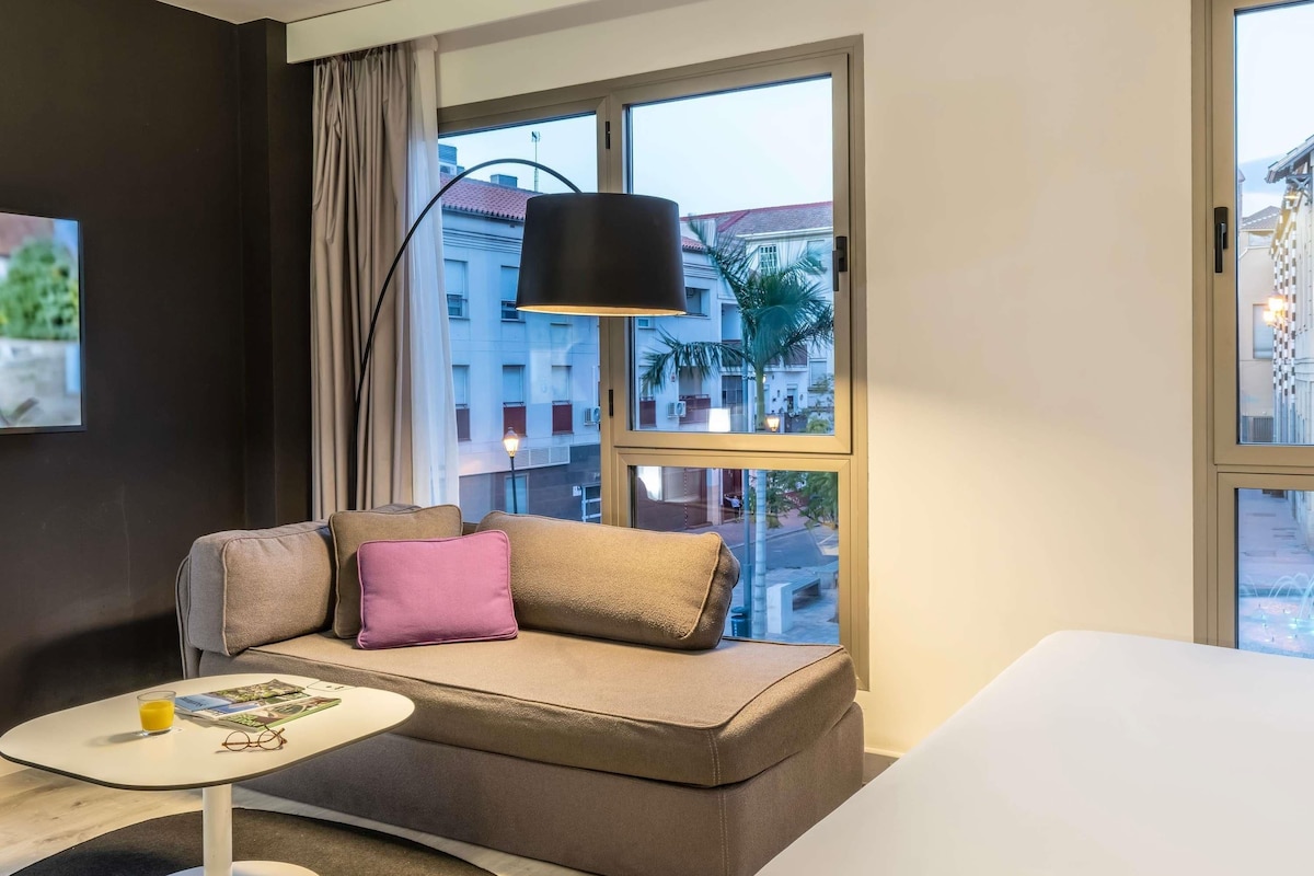 A modern hotel room with a sofa, a small table, and a large window with a view of buildings.