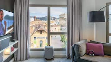 A hotel room with a large window, a television, a sofa, a small table with a drink, and a suitcase.