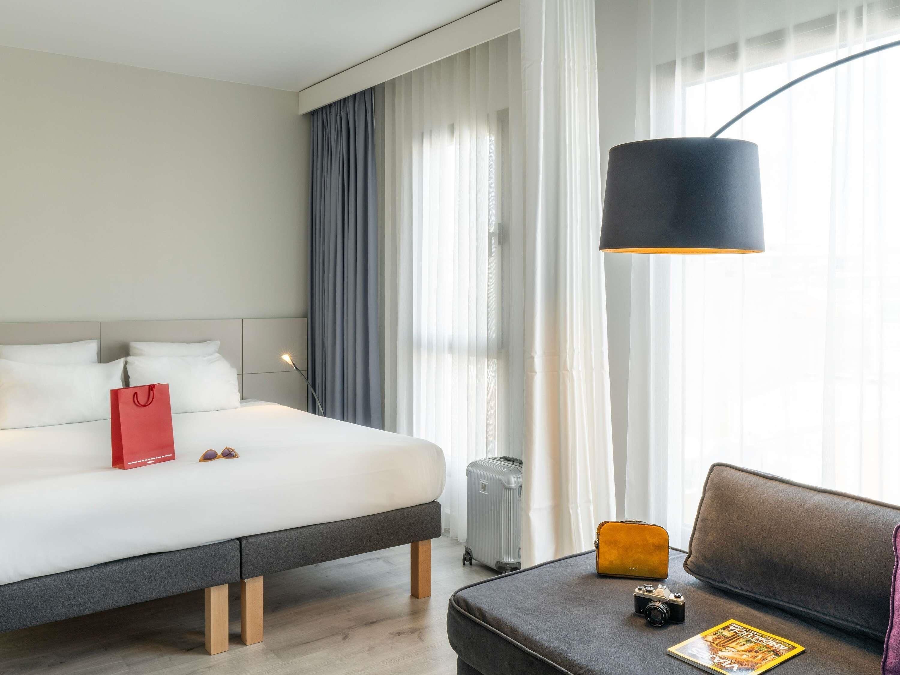 A hotel room with a bed, a bedside table with a lamp, a suitcase, a small table with a camera and a magazine, and a couch.