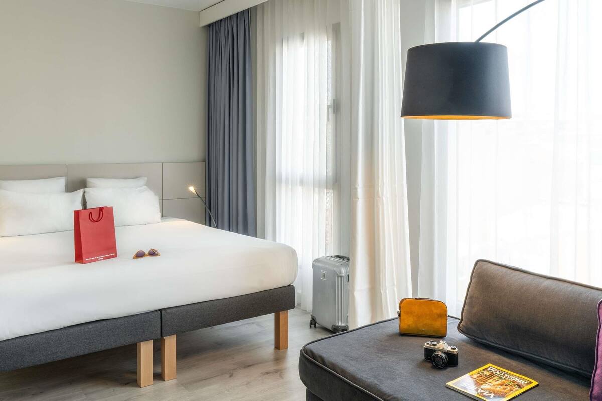 A hotel room with a bed, a bedside table with a lamp, a suitcase, a small table with a camera and a magazine, and a couch.